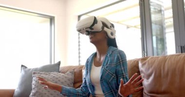 Genç bir Afrikalı Amerikalı kadın evdeki bir kanepede kulaklıkla sanal gerçekliği keşfediyor. VR kulaklığı takıyor, elleriyle işaret ediyor ve yavaş çekimde kendini kaptırmış gibi görünüyor.