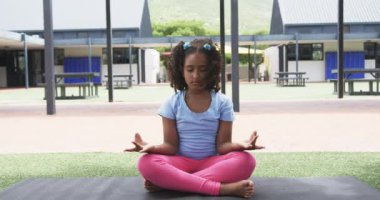 Çift ırklı kız, okul bahçesinde gözleri kapalı meditasyon yapar. Mavi gömlek ve pembe pantolon giyer. Farkındalık egzersizi sırasında sakin olur. Yavaş çekim..