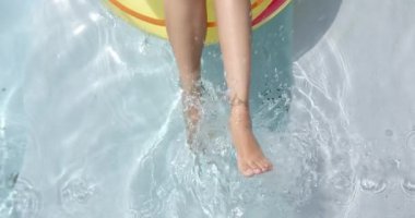 Şişme bir yüzükten havuzda sarkan genç bir kadının bacaklarının yakın çekimi. Temiz su ve parlak güneş ışığı havuzda sakin bir gün geçirdiğimizi gösteriyor..