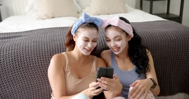 2人の若い女性の友人は 顔のマスクとヘッドバンドを持つスマートフォンで笑っている 彼らは居心地の良い部屋でベッドに座ってスマートフォンを見ながら一緒に笑っている — ストック動画