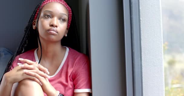 一位年轻的非洲裔美国妇女沉思地凝视着窗外 家里有复制的空间 她的辫子上有红光 头戴宽松的红色上衣 动作缓慢 — 图库视频影像