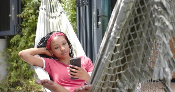 一位年轻的非洲裔美国妇女用智能手机躺在后院的吊床上 她把头发剪成红色 对着智能手机笑 穿着休闲装 动作缓慢 显得很放松 — 图库视频影像