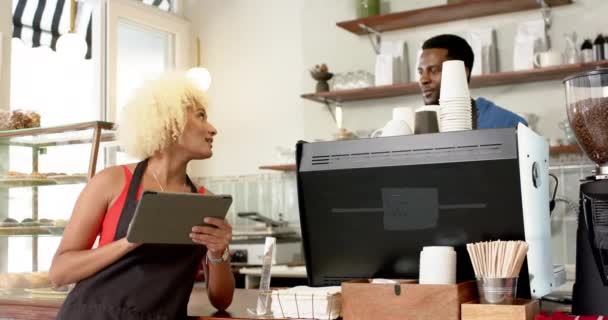 一位年轻的非洲裔美国男子在一家咖啡店用石碑和一位黑人妇女一起工作 她拿着平板电脑 对着他笑着 这表明她在随意的工作环境中和他进行了友好的互动 动作缓慢 与快乐的咖啡师团队一起工作 — 图库视频影像