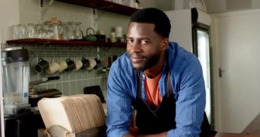 Genç bir Afro-Amerikalı erkek barmen kafeteryaya yaslanmış sıcak bir şekilde gülümsüyor. Turuncu bir tişörtün üzerine mavi bir gömlek giyer. Ev ortamında rahat eder, yavaş çekimde..
