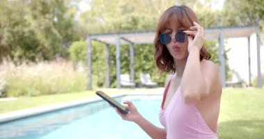 Pembe mayo giymiş melez bir kadın havuz kenarında fotokopi çekerken gülümsüyor. Açık havada güneşli bir günün tadını çıkarıyor. Kahverengi saçları ve güneş gözlükleri rahat bir havayı yansıtıyor..
