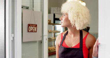 Kıvırcık sarı saçlı melez bir kadın açık bir kafe kapısında duruyor. Kırmızı ve siyah bir önlük giyiyor. Bu da bir dükkanda ya da kafede çalıştığını gösteriyor.. 