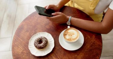 Kafe masasında elinde donut ve kafede kahve olan akıllı telefon kullanan melez bir kadın. Sarı bir bluz giyer, telefonunu karıştırırken atıştırmayı sever, sıradan bir anı gösterir..