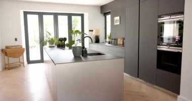 Modern bir mutfakta parlak gri dolaplar ve lavabosu olan merkezi bir ada bulunur. Doğal ışık alanı büyük cam kapılardan sarıyor, temiz ve minimalist tasarımı vurguluyor..