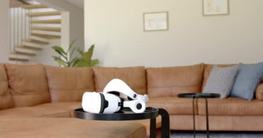 VR kulaklığı sehpanın üzerinde duruyor. Oturma odası büyük bir kanepe ve duvarları süsleyen ahşap çerçevelerle dolu, ağır çekimde.