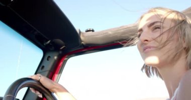 Yolculukta araba kullanan beyaz saçlı genç bir kadın. Neşeli yüzünü aydınlatan güneş ışığı. Rahat ve mutlu görünüyor. Arkasında açık mavi bir gökyüzü olan bir aracı kullanıyor..