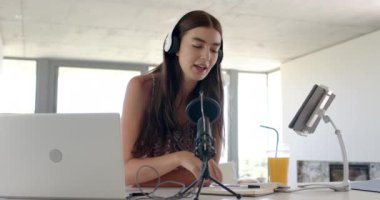 Uzun kahverengi saçlı beyaz bir kız bir podcast kaydediyor. Kulaklık takıyor, mikrofona konuşuyor, önünde dizüstü bilgisayar ve not defteri var, ağır çekimde..