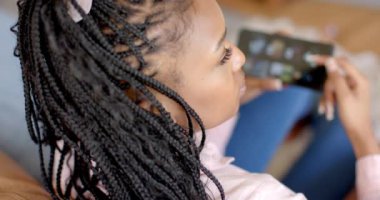 Genç bir Afrikalı Amerikalı kadın evdeki kanepede otururken akıllı telefonuna odaklanıyor. Saçlarını ördü, göze çarpmayan bir makyajı var, ve günlük giyinmiş, biraz müzik dinlemekten hoşlanıyor, yavaş çekim..