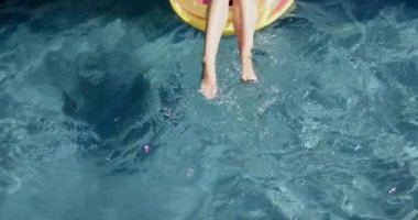 Genç bir kızın bacakları görünür, açık mavi havuz suyuna asılı, fotokopi alanı olan sarı bir şamandırada. Güneş ışığı sudan yansıyor, havuzda sakin bir gün olduğunu gösteriyor, ağır çekim..