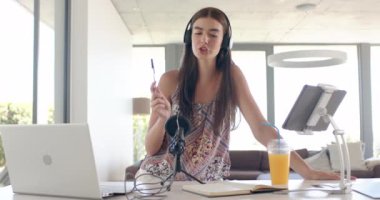 Uzun kahverengi saçlı beyaz bir kız dizüstü bilgisayar ve kulaklıkla podcast kaydediyor. Modern bir ofis ortamında, bir çalışma ya da çalışma sırasında neşeli bir anı öneriyor..