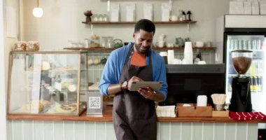 Genç bir Afrikalı Amerikalı erkek barista elinde tabletle bir kafe tezgahının arkasında duruyor. Mavi gömleğinin üzerine kahverengi bir önlük giyiyor. Profesyonel ve misafirperver bir tavır sergiliyor. Ağır çekim..