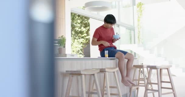 Teini Ikäinen Aasialainen Poika Punaisessa Paidassa Istuu Keittiön Pöydällä Syö tekijänoikeusvapaata kuvapankin filmiä