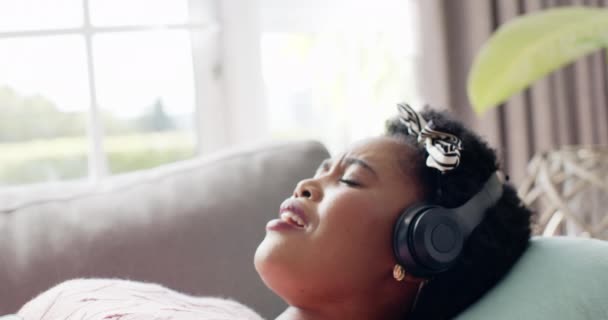 アフリカ系アメリカ人女性はヘッドフォンで音楽を楽しんでおり 目を閉じて自宅で音楽を楽しんでいる 彼女はソファーでリクライニングし 明るいリビングルームで喜びとリラクゼーションを表現しています — ストック動画