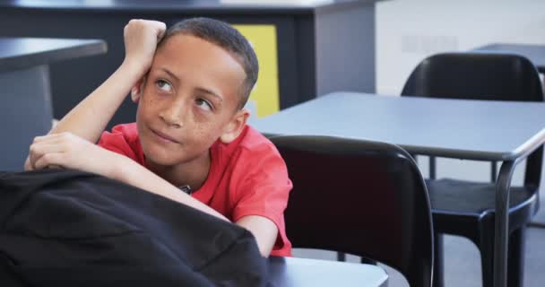 一个长雀斑的小孩坐在学校的教室里 心事重重 他穿着一件红色的衬衫 把头靠在手上 周围都是桌子 动作缓慢 — 图库视频影像