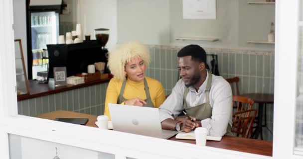若いアフリカ系アメリカ人男性のバリスタチームと カフェでラップトップについて話し合う女性 彼は灰色のエプロンを着て 彼女が黄色いトップにいる間 バリスタであるかもしれないことを示唆し 会話に従事 — ストック動画