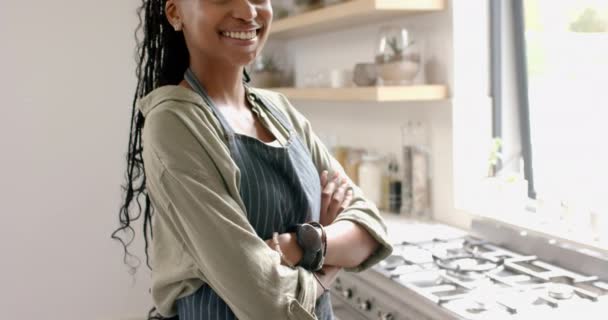 アフリカ系アメリカ人の若い女性が自宅の明るいキッチンに立っています 彼女は長い編み毛 輝く笑顔を持っており エプロンでカジュアルなシャツを着ています スローモーション — ストック動画