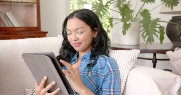 一个黑头发的年轻的黑人妇女在沙发上使用平板电脑 她穿着一件蓝色格子呢衬衫 在舒适的家庭环境中显得很放松 很惬意 动作缓慢 — 图库视频影像