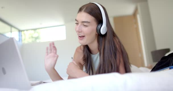 ヘッドフォンが付いている十代の白人少女は自宅で彼女のラップトップのビデオ通話に従事しています 彼女はベッドの上に横たわり 会話中にジェスチャーし カジュアルでリラックスした設定を示唆しています — ストック動画