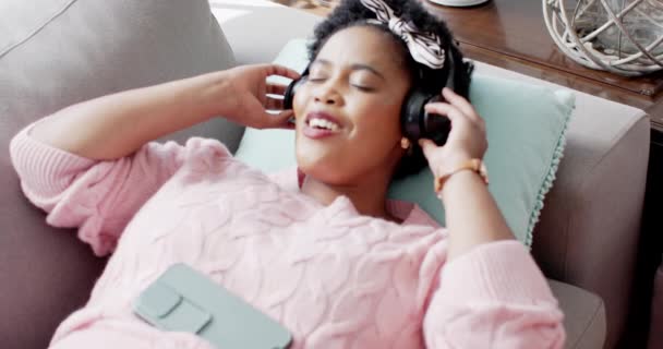 非洲裔美国妇女喜欢在家里的沙发上听音乐 闭上眼睛 她沉浸在声音中 穿着粉色毛衣 戴着黑色耳机 动作缓慢 — 图库视频影像