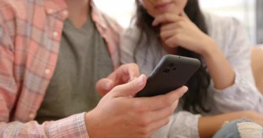 Çeşitli bir çift evdeki koltukta akıllı telefon kullanıyor. İspanyol kadın ekranı gösterirken Asyalı adam aleti tutuyor, hem ilgi gösteriyor, hem de değişmemiş, yavaş çekim..