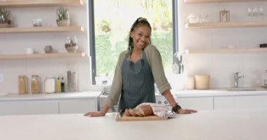 Genç bir Afrikalı Amerikalı kadın taze pişmiş ekmekle modern bir mutfakta duruyor. Saçları örgülü, ışıltılı bir gülüşü var ve sıradan bir kıyafetin üzerine kot önlük giyiyor, ağır çekimde, değiştirilmemiş..