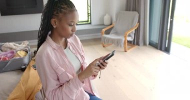 Genç bir Afrikalı Amerikalı kadın akıllı telefonuna odaklanmış, kopyalama alanı olan. Saçlarını örüyor, pembe bir gömlek giyiyor, ve evde oturuyor, kişisel eşyalarıyla çevrili, ağır çekimde, değiştirilmemiş..
