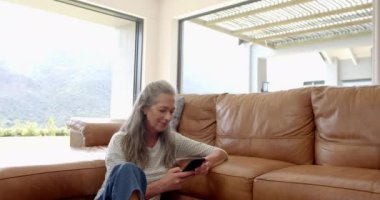 Olgun, beyaz bir kadın evdeki bir kanepede telefonunun fotokopisini çekiyor. Uzun gri saçları, minimum makyajı var ve bronz deri koltukta rahat bir şekilde oturuyor, ağır çekimde, değiştirilmemiş..