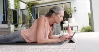 Yetişkin bir Kafkas kadın evde antrenman yaparken tablet kullanıyor. Gri saçları var, günlük spor kıyafetleri giyiyor ve yoga minderinin üstünde, ağır çekimde, değişmemiş..