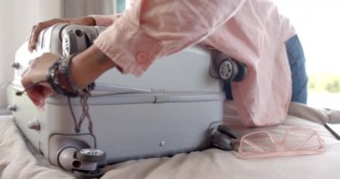 Genç bir Afrikalı Amerikalı kadın tatil için gümüş bir bavul hazırlıyor. Yakınlarda akıllı telefonu ve pembe güneş gözlüğü var. Seyahat hazırlıkları, yavaş çekim, değişmemiş..