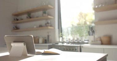 Bir tablet ve defter mutfak tezgahında duruyor. Güneş ışığı pencerelerden süzülüyor, açık raflı ve sakin bir atmosferi olan modern bir mutfağı vurguluyor, yavaş çekim, değiştirilmemiş.
