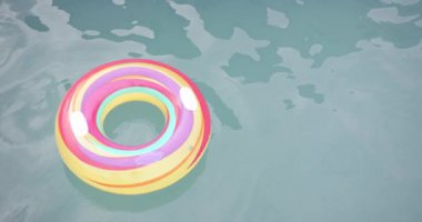 Renkli bir şişme halka havuzun yüzeyinde yüzer. Güneş ışığı sudan yansıyarak, halkanın canlı renklerini ve hafif dalgalanmalarını vurgular, değişmez, yavaş çekim