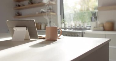 Güneşli bir mutfakta fotokopi odası olan bir tabletin yanında dumanı tüten bir kupa. Sıcak güneş ışığı odayı yıkıyor, eldeki teknolojiyle huzurlu bir sabahı vurguluyor, ağır çekimde, değişmeden..