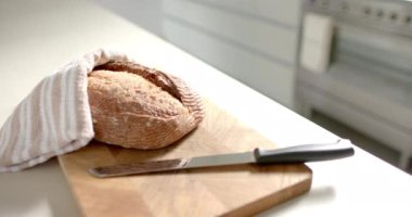 Tahtanın üzerinde dilimlenmeye hazır bir somun ekmek duruyor. Yakınlarda bir bıçak duruyor, mutfak tezgahı temiz, parlak bir zemin sağlıyor, yavaş çekim, değiştirilmemiş..