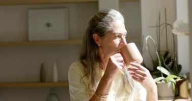 Olgun, beyaz bir kadın evinde sıcak bir içecek içiyor. Uzun gri saçları ve düşünceli bakışlarıyla, gündelik giyinmiş, sakin bir anın tadını çıkarıyor, ağır çekim, değişmemiş..