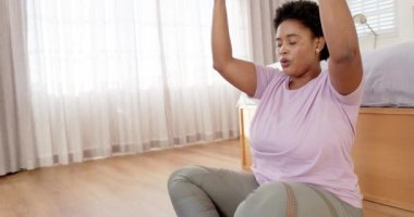 Afro-Amerikalı kadın sakin bir odada meditasyon yapıyor, arka planda bej perdeler ve fotokopi alanı var. Sıradan bir kıyafet ve akıllı bir kaset giyer, nefesine odaklanır, değişmez, ağır çekimde..