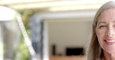 Olgun, beyaz bir kadın geniş bir gülümseme sergiliyor. Uzun, gri çizgili sarı saçları, gülme çizgileri ve doğal bir makyajı var. Ağır çekimde, değişmemiş..