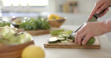 Elleri ahşap bir tahtanın üzerinde salatalık dilimliyor, mutfak düzeninde. Olgun beyaz kadın salata hazırlıyor, sağlıklı bir yaşam tarzı sergiliyor, ağır çekimde, değiştirilmemiş..