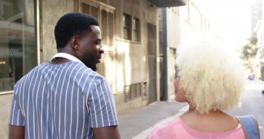 Çeşitli bir çift şehir tatili için şehir sokaklarında geziniyor. Afro-Amerikalı adam geriye bakar, çizgili bir gömlek spor, kıvırcık sarı saçlı beyaz kadın ileriye bakar, pembe bir bluz, değiştirilmemiş