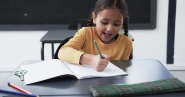 Okul ortamında, melez bir kız yazmaya odaklanır. Kahverengi saçlı, sarı bir bluz giyiyor ve işinde yeşil kalem kullanıyor, değiştirilmemiş, ağır çekimde.