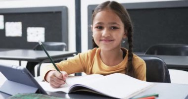 Bir okul ortamında, genç beyaz bir kız bir deftere yazı yazıyor. Örgülü uzun kahverengi saçları, sarı bir bluzu var, ve masanın üzerinde bir tablet kullanıyor, değiştirilmemiş, ağır çekimde.