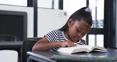 Bir okul sınıfında, melez bir genç kız bir kitap okumaya odaklanır. Siyah saçlı, mor bir aksesuarla bağlanmış ve çizgili, değiştirilmemiş ağır çekimde bir bluz giyiyor.