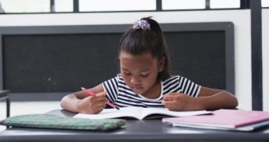 Bir okul sınıfında, melez bir kız ders kitabına konsantre olur. Saçları mor bir aksesuarla bağlanmış ve üzerinde çizgili bir kıyafet var..