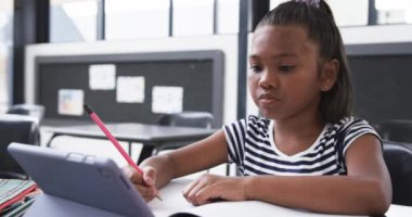 Bir okul sınıfında, melez bir kız tabletine odaklanıyor. Uzun, siyah saçlı, çizgili bir bluz giyiyor, ve bir deftere yazıyor, değiştirilmemiş, yavaş çekim..