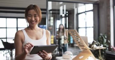 Orta yaşlı bir Asyalı üst düzey yönetici sıradan bir iş ofisinde tabletine odaklanır. Omuz hizasında saçları, yeşil bluzu var. Modern bir ofiste, tuğladan bir duvarı var. Değiştirilmemiş, ağır çekim..