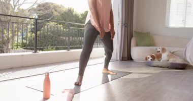 Yaşlı bir Afrikalı Amerikalı kadın evde yoga yapıyor. Kısa gri saçları var, pembe kolsuz bluz giyiyor ve köpeği yatağının yanında duruyor, değişmemiş, ağır çekimde.
