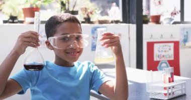 Okulda, sınıfta, genç bir Afrikalı Amerikalı öğrenci kimyasal mataraları inceliyor. Meraklı bir ifadesi var. Güvenlik gözlüğü ve açık mavi bir tişört giyiyor. Değiştirilmemiş, ağır çekim..